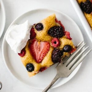 A slice of berry sheet cake on a white plate with fork and dollop of cream.