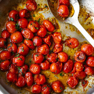 Blistered tomatoes in a metal pan with a large spoon.