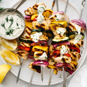 Several Greek chicken kabobs on a plate with lemon wedges and tzatziki sauce.